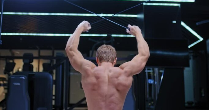 Athlete, a bodybuilder, stretches the harnesses behind the head on the simulator, trains the latissimus dorsi. Embossed inflated back muscles. View from the back. 4k, ProRes