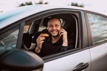 Portrait of a smiling young caucasian businessman sitting in a car and talking on the phone and holding a donut with chocolate in his hand