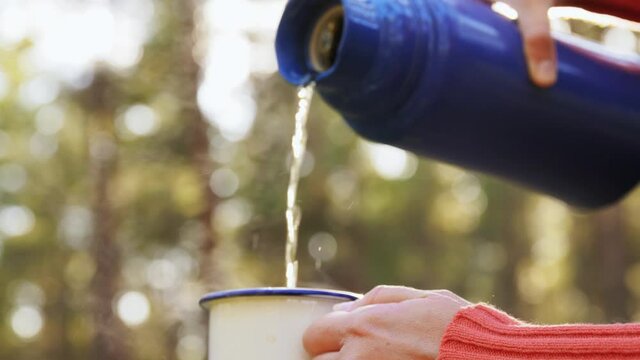 drinks, leisure and people concept - close up of hands pouring tea from thermos to mug in forest