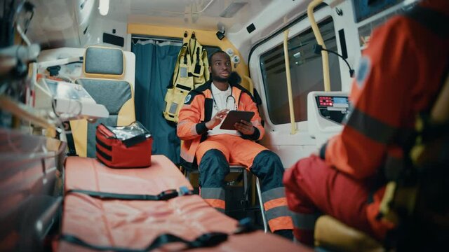 Black African American Paramedic Using Tablet Computer while Riding in an Ambulance Vehicle for an Emergency. Emergency Medical Technicians are on Their Way to a Call Outside the Healthcare Hospital.