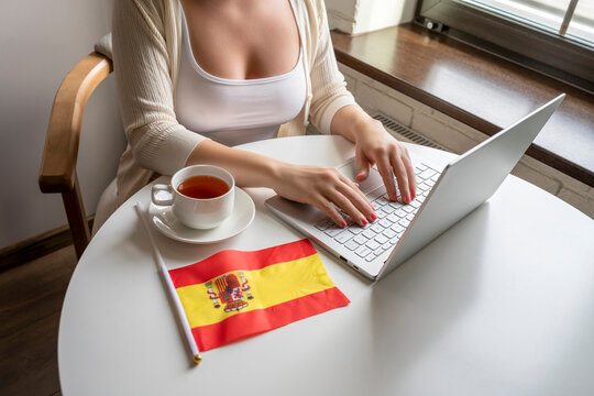 Lonely Woman Freelancer With Flag Of Spain Enjoying Having Breakfast With Cup Of Coffee Working On Laptop Sitting Near Window In Cafe At Morning.