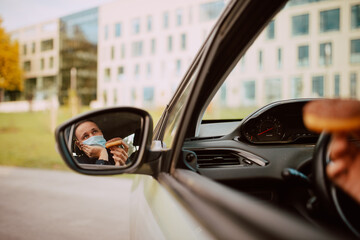 A reflection of a sad man with a medical face mask in the rear view mirror while sitting in a car with a donut with chocolate in his hand. Meal in the car during the pandemic COVID - 19 coronavirus