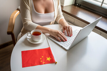 Lonely woman freelancer with flag of China enjoying having breakfast with cup of coffee working on laptop sitting near window in cafe at morning.
