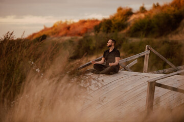 A beautiful young caucasian man sitting on an old bridge, meditating and looking away, while there is tall grass around him in the frame. Yoga and meditation in the fresh mountain air