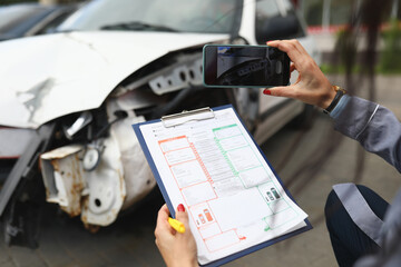 Woman photographs a broken car on smartphone and holds insurance documents in her hands. Damage...