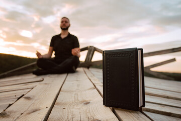 The black book stands on the bridge in focus, and in the background is a young caucasian man meditating. Yoga on the mountain