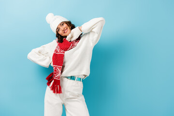 pleased young woman in white winter outfit, warm scarf, gloves and hat on blue