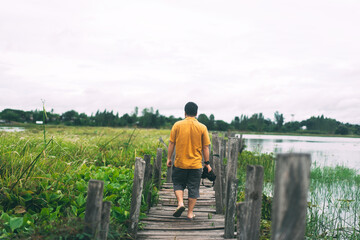 Rear view of adult camera travel man relax walking over old wooden bridge at lake.