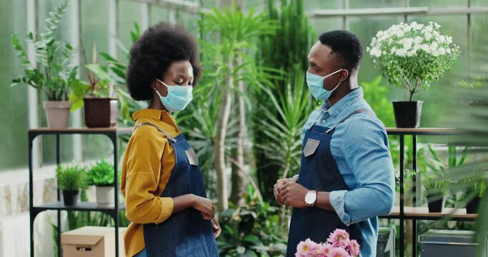 Portrait of African American male and female workers greeting with elbows in floral center at work. Cheerful woman and man florists in masks talking in good mood. Employment concept