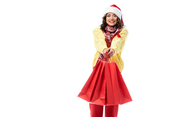 joyful young woman in santa hat and scarf with ornament holding red shopping bags isolated on white