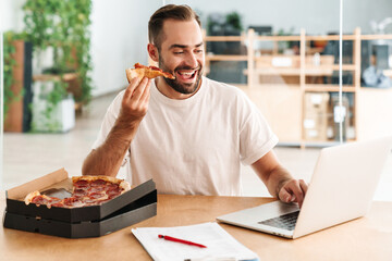 Caucasian joyful man smiling and eating pizza while working with laptop