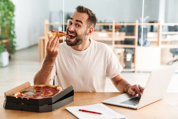 Caucasian joyful man smiling and eating pizza while working with laptop