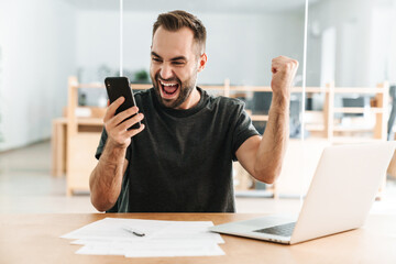 Man making winner gesture and using cellphone while working with laptop
