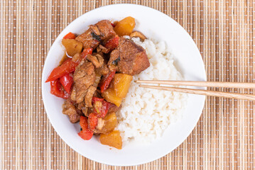 Adobo pork with pineapple and pepper in a bowl of boiled rice and chopsticks on a bamboo napkin, close up, top view