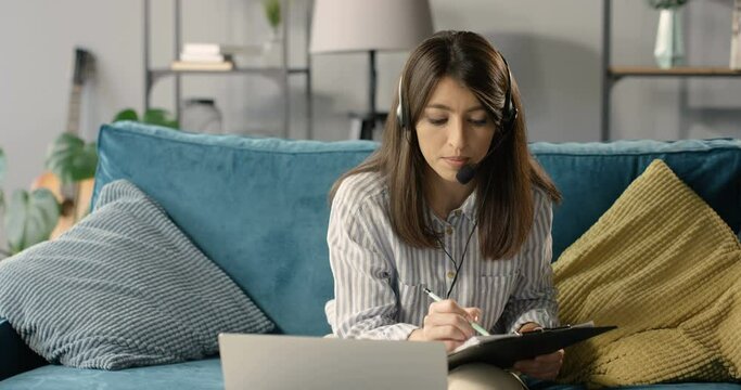 Businesswoman in headset and mic sitting on couch, videochating on laptop computer and noting information. Female teacher or cauch teaching online. Freelancer working at home via webcam with videochat