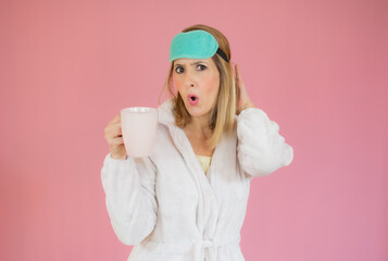 Young woman wearing pajama and sleep mask drinking coffee over isolated pink background.