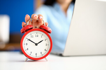 Woman's hand lies on red alarm clock next to laptop. Effective use of working time concept