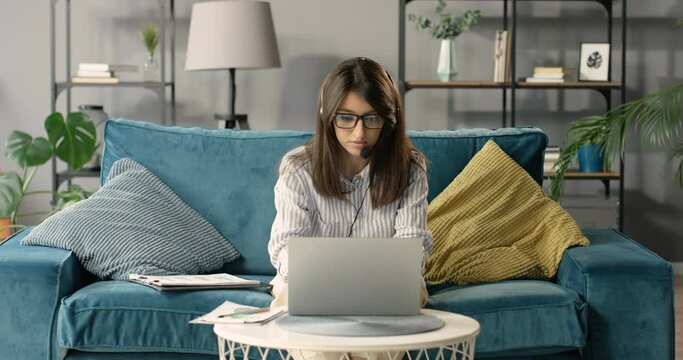 Beautiful Caucasian businesswoman in headset sitting on couch, typing laptop computer and working. Female teacher or cauch work online. Freelancer doing job at home via webcam on sofa in cozy room.