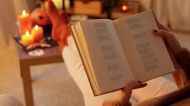 halloween, holidays and leisure concept - young woman reading book and resting her feet on table at cozy home