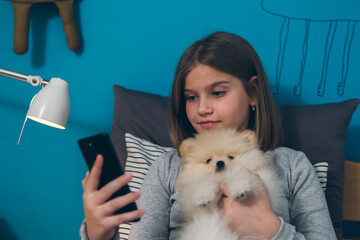 girl sitting on bed with her puppy in her room and using mobile phone