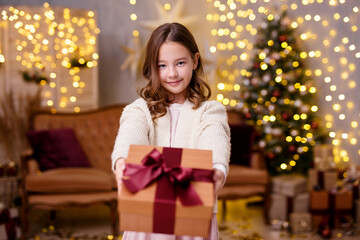 Fototapeta na wymiar Christmas and new year concept - cute little girl giving gift box in decorated living room with Christmas tree and lights