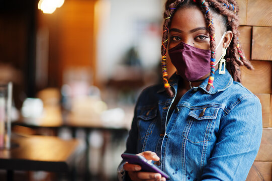 Stylish african american woman with dreadlocks afro hair, wear jeans jacket and face protect  mask at restaurant, hold cellphone. New normal life after coronavirus epidemic.
