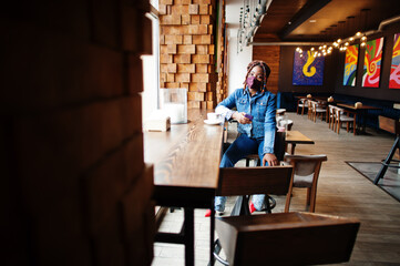 Fototapeta na wymiar Stylish african american woman with dreadlocks afro hair, wear jeans jacket and face protect mask at restaurant, hold cellphone. New normal life after coronavirus epidemic.