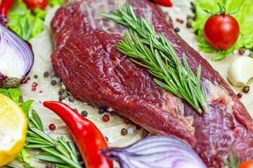 Raw red beef meat with green rosemary and fresh vegetables on light wooden cutting board background.
