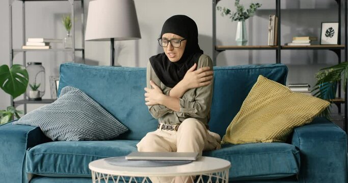 Yong Arabian woman in glasses and hijab sitting on couch and having symptoms of cold, flu or coronavirus. Female in traditional headscarf caughing, sneezing and having fever with high temperature.