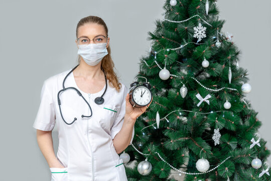 A Female Doctor In A Protective Medical Mask Holds An Alarm Clock On The Background Of A Christmas Tree. Timely Access To Medical Care During The Christmas Holidays And New Year