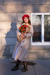 Full length vertical portrait of beautiful blue-eyed smiling young woman wearing a trench coat and red beret standing on sidewalk in dappled light holding baguettes and tulips