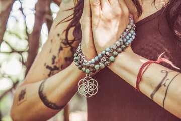 Female hands doing namaste mudra yoga practice in forest with mala necklace - obrazy, fototapety, plakaty