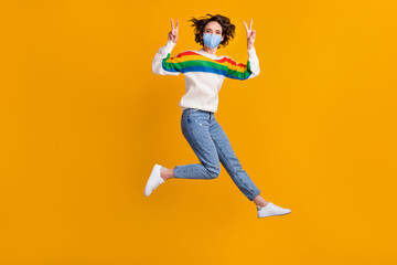 Full size photo of funny woman jump show v-sign wear mask sweater jeans footwear isolated on yellow background