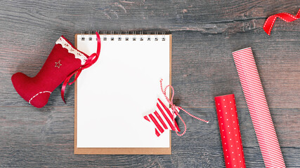 Fototapeta premium Merry Christmas mockup. Christmas stocking an open notepad on wooden background, red gift wrapping paper. New Year season greetings card resolutions mockup or goals. flat lay copy space