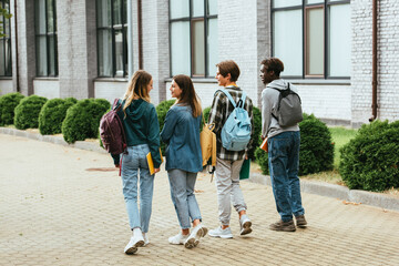 Fototapeta premium Back view of smiling teenagers with backpacks walking on urban street