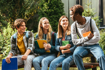 Smiling teenagers with books looking at african american friend on bench outdoors