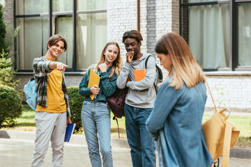 Fototapeta premium Selective focus of smiling multicultural teenagers with backpacks pointing at sad girl outdoors