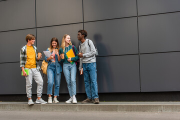 Fototapeta premium Cheerful multicultural teenagers with books and coffee to go standing near building outdoors