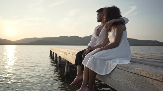 Romance scene of heterosexual mixed race couple looking at the setting sun sitting on a pier in the lake - Caucasian fine man embracing her black Hispanic curly girlfriend in a loving gesture