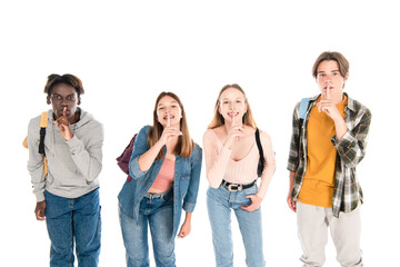Smiling multiethnic teenagers with backpacks showing shh gesture isolated on white