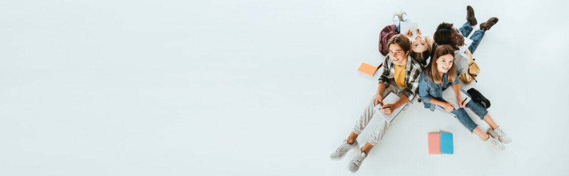 Panoramic Shot Of Smiling Multicultural Teenagers Smiling While Writing On Notebooks On Grey Background