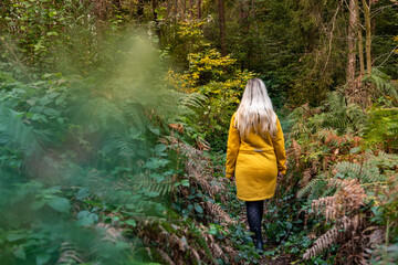 Blonde woman in yellow jacket walking along a forest path 	