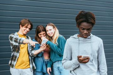 Selective focus of african american boy using smartphone near laughing teenagers outdoors