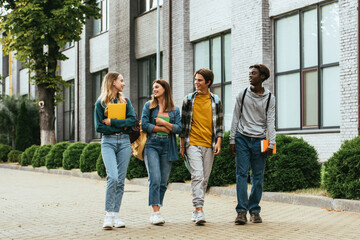 Fototapeta premium Smiling multicultural teenagers with notebooks and backpacks walking on urban street