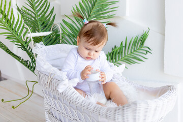 cute little baby girl sitting in a beautiful stroller in a white bodysuit at home and holding a...