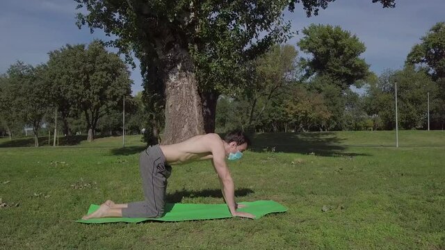 Young fit man with the mask doing yoga on a mat in the park, attractive confident man doing yoga cat and cow pose