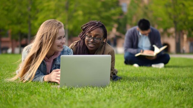 Diverse friends students lying outdoors on lawn with laptop chatting and smiling
