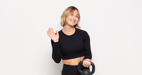 young blonde woman smiling happily and cheerfully, waving hand, welcoming and greeting you, or...