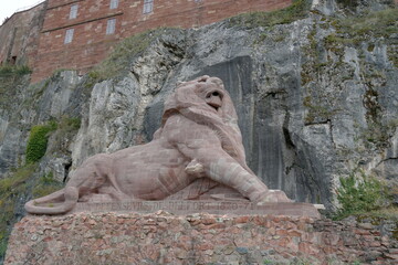 Fototapeta na wymiar A statue of a lion made of pink sandstone in the fortress or citadel in Belfort, France. On the background there is a rock wall in contrasting color. On the top of it there is building of the citadel.