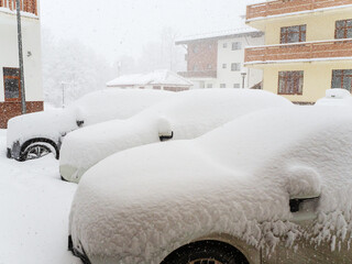 Parked cars strewn with snow against the background of houses during a blizzard